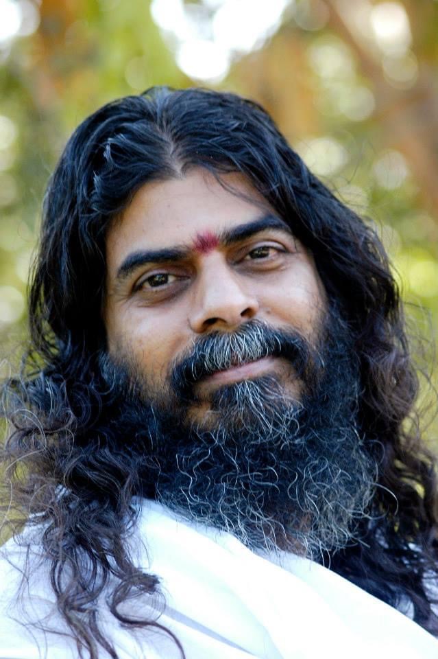 Shri Nandanji Swatantra, founder of Vanaprastham Charitable Trust: a man with long black hair and a grey-streaked beard, a red tilak on his forehead and a white shawl over his shoulders, looking calmly at the camera against a softly blurred background of sunlit foliage.