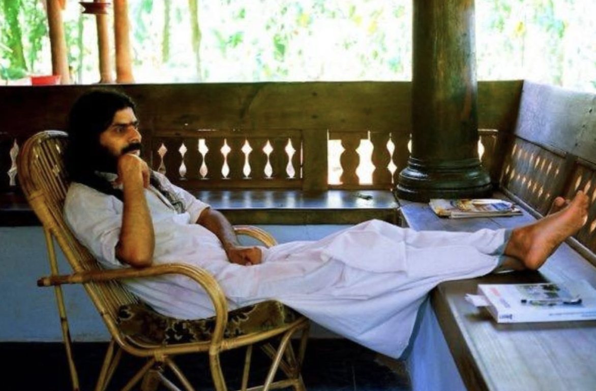 Shri Nandanji, founder of Vanaprastham Charitable Trust, seated in a cane armchair on a wooden Kerala veranda: dressed in white, bare feet resting on the bench ledge and one hand at his chin, carved wooden railings and sunlit foliage behind him.